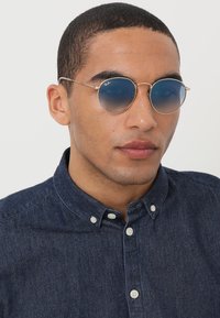 Occhiali da sole rotondi con lenti blu sfumate e montatura in metallo dorato. Chi li indossa ha una camicia in denim scuro con bottoni.