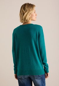 Grönblå ulltröja med ribbad rund halsringning, långa ärmar och en något längre bakre fåll. Tyget har ett mjukt, texturerat utseende.