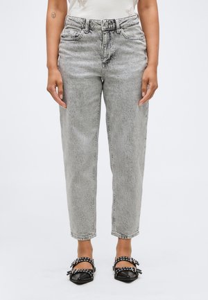 Femeie care poartă blugi gri cu spălare acidă, talie înaltă, croială lejeră și pantofi negri cu vârf ascuțit, cu barete argintii cu găici.