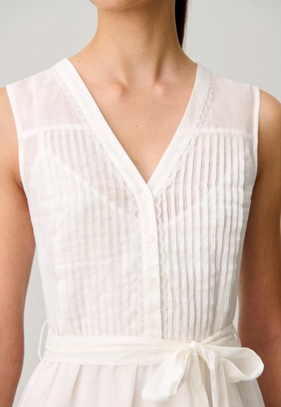 Vestito bianco senza maniche con pieghe verticali, davanti con bottoni, delicato bordo di pizzo lungo la scollatura e cintura in tessuto annodata a fiocco in vita.