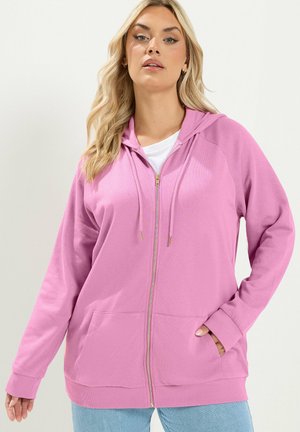 Frau mit langen blonden Haaren, die einen rosa Zip-Hoodie über einem weißen Shirt und hellblauen Jeans trägt und mit einer Hand in der Tasche posiert.