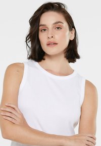 White sleeveless top with a ribbed texture, round neckline, and minimalist design. Model displays arms crossed with a subtle tattoo on shoulder.