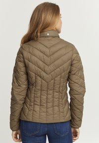 Khaki puffer bunda s diamantovým prešívaním, vysokým golierom a detailmi na patentkách, v kombinácii s modrými džínsami.