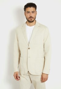 Blazer beige de tejido ligero, con un solo botón, dos bolsillos delanteros y un corte entallado, llevado sobre una camiseta blanca.