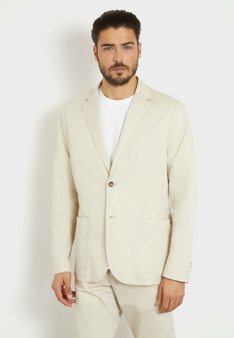 Blazer beige de tejido ligero, con un solo botón, dos bolsillos delanteros y un corte entallado, llevado sobre una camiseta blanca.