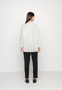 Camisa branca de manga longa com colarinho, punhos abotoados e um corte descontraído, combinada com calças pretas ajustadas e sapatos pretos.
