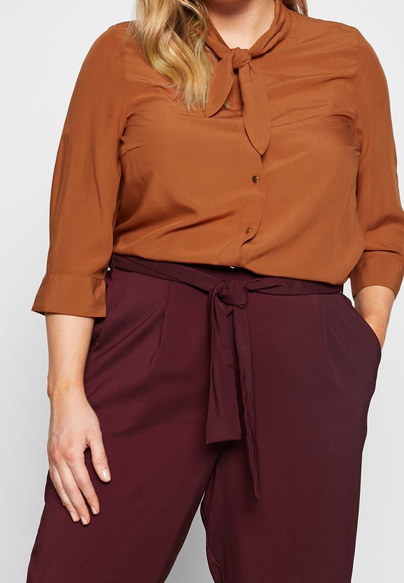 Femme portant un chemisier couleur rouille avec un col à nouer et des manches trois-quarts, associé à un pantalon taille haute bordeaux foncé avec une ceinture en tissu.