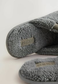 Close-up of soft, gray, fuzzy slippers with fabric tags labeled "NEXT" on the inner sole, on a light-colored surface.