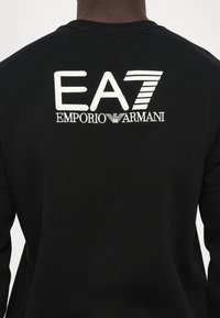 Črn dukserica z velikim vezenim logotipom EA7 in besedilom "EMPORIO ARMANI" na hrbtu, z gladko teksturo tkanine.