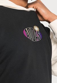 Černá mikina s bílou kapucí, s kulatým logem Adidas v fialové, černé a zlaté barvě. Tkanina má hladkou strukturu.