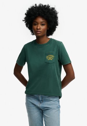 Mujer con cabello afro que lleva una camiseta verde oscuro de Superdry y vaqueros azul claro, de pie con las manos detrás de la espalda frente a un fondo blanco.