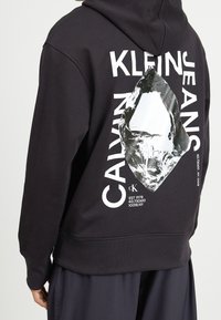 Černá mikina s kapucí, kterou nosí osoba, s bílým nápisem Calvin Klein Jeans a na zádech reflexivním stříbrným abstraktním motivem.