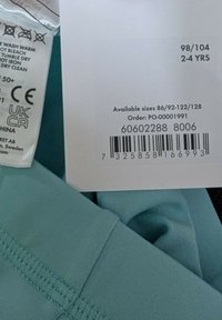 Een turquoise stoffen label met wasinstructies en maatinformatie, met gedrukte details en een streepjescode op een zacht materiaal.