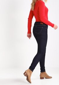 Jeans skinny de denim azul oscuro con cintura alta, cuentan con bolsillos traseros y un parche de cuero marrón en la cintura, combinados con botas de tobillo beige.