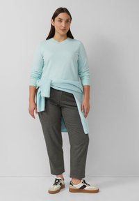 Vrouw in een lichtblauwe trui en een bijpassende geknoopte trui om haar taille, grijze broek en witte sneakers met zwarte details staat tegen een grijze muur.