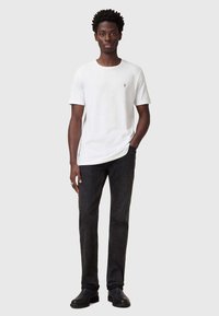 Hvid T-shirt med korte ærmer og et lille broderet logo, parret med mørke denimjeans og sorte ankelstøvler. Ensfarvet baggrund.
