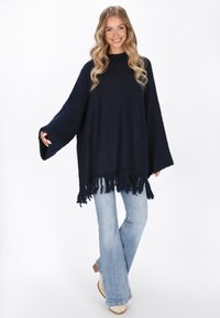 Navyblauer oversized Poncho-Pullover aus strukturiertem Stoff, mit hohem Kragen und fransigem Saum. Kombiniert mit hellblauen Schlagjeans.