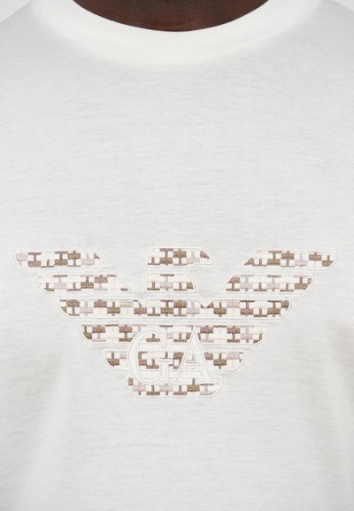 Balts kokvilnas T-krekls ar lielu bēša un brūnu izšuvumu logotipu ar spārniem un "GA" centrā. Gluda tekstūra.