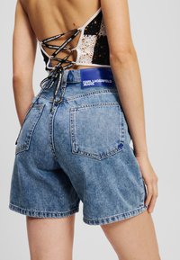 Jeans kratke hlače srednje modre barve z visokim pasom, raztrganimi roba in zadnjimi žepi. Imajo sijoč modri logotip na hrbtu.