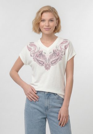 Femme blonde aux cheveux courts portant une chemise blanche à col en V avec des motifs cachemire roses, associée à un jean en denim bleu clair.