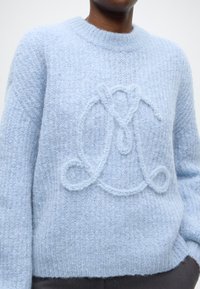 Pull en maille bleu clair avec un col rond. Il présente un tissu texturé, des poignets côtelés et un motif logo en relief sur le devant.