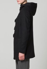 Cappotto in lana nera con cappuccio, chiusure a toggle, tasche laterali e una silhouette sagomata, che mette in risalto una texture liscia e linee pulite.