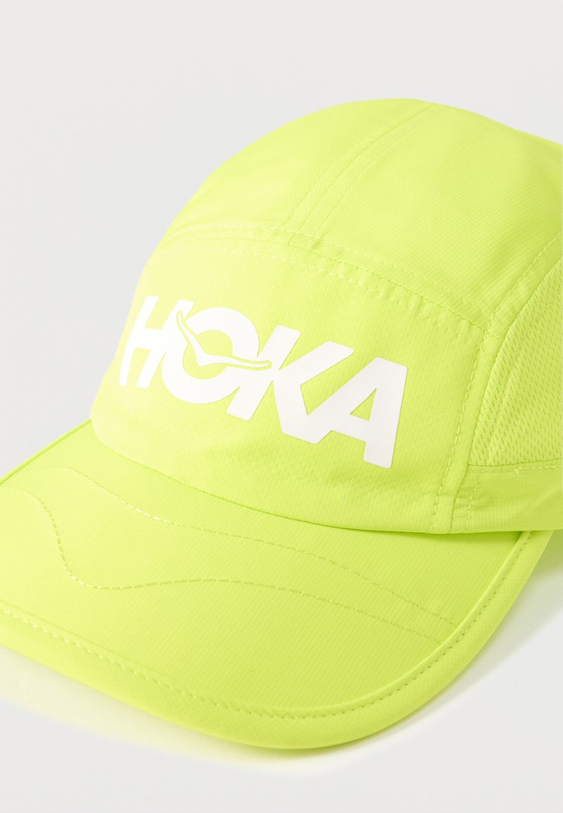Gorra deportiva de color amarillo-verde brillante con el logo blanco "HOKA" y paneles de malla a los lados, colocada sobre una superficie blanca.