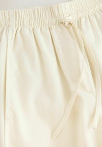 Ceinture élastique couleur crème avec cordon de serrage, texture lisse et surfaces avant plates ; présente des coutures décoratives et des poches latérales.