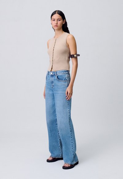 Top senza maniche beige in maglia con bottoni dorati, abbinato a jeans ampi di colore azzurro chiaro. Il modello indossa sandali scuri e un accessorio per il braccio di colore marrone.