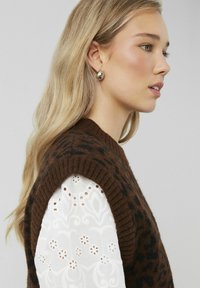 Un gilet en laine marron avec des taches de léopard noires porté par-dessus une chemise à manches longues en dentelle blanche affichant un motif floral. Boucles d'oreilles rondes en argent.