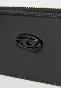 Black patent leather pouch featuring a zip closure and a prominent black oval logo accent. Smooth surface with subtle stitching detail.