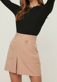 Mujer vestida con una mini falda beige con grandes bolsillos frontales y un detalle de anillo de metal, combinada con una blusa negra de manga larga y un collar de cadena plateada.