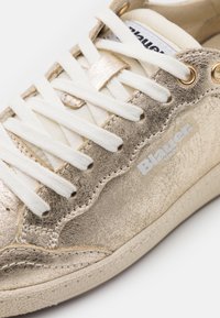 Baskets dorées métalliques avec finition texturée, lacets blancs et œillets dorés. Présentent un design courbé et un détail de logo sur le côté.