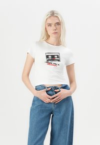Balts īss t-krekls ar kasetes grafiku un tekstu "Es tev izveidoju mixtape", kombinēts ar augsto jostasvietu ziliem džinsiem.