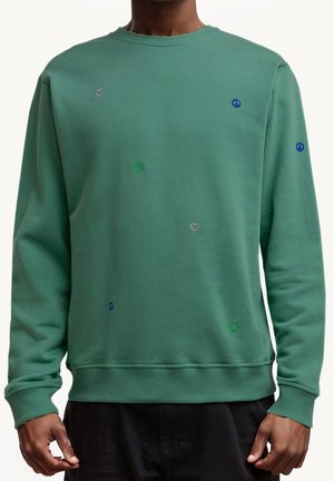 Mann trägt grünes Sweatshirt mit kleinen, grünen, bestickten Smiley-Gesichtern, blauen Peace-Zeichen und silbernen Herzen, steht vor weißem Hintergrund.