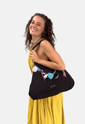 Jonge vrouw die glimlacht, gekleed in een gele jurk, met een grote zwarte tas waarin kleurrijke sleutelhangers en accessoires aan de schouderband hangen.