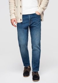 Jean en denim bleu avec une coupe classique droite, une décoloration subtile et un design à cinq poches. Accompagné d'un cardigan en maille beige clair et de chaussures foncées.