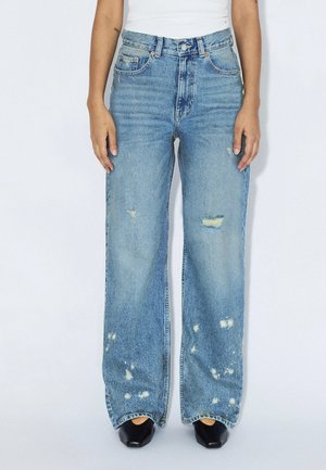 Jean droit bleu avec des patchs usés, porté avec des chaussures noires pointues et un haut blanc, montré de la taille aux pieds.