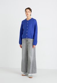 Blue knitted cardigan with long sleeves and a round neckline, featuring buttons down the front. Paired with wide-leg grey jeans and grey sneakers.