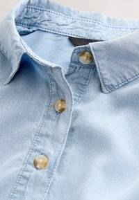 Camisa de mezclilla azul claro con cuello clásico, dos botones transparentes y detalles de costuras visibles. Textura suave con un diseño casual.