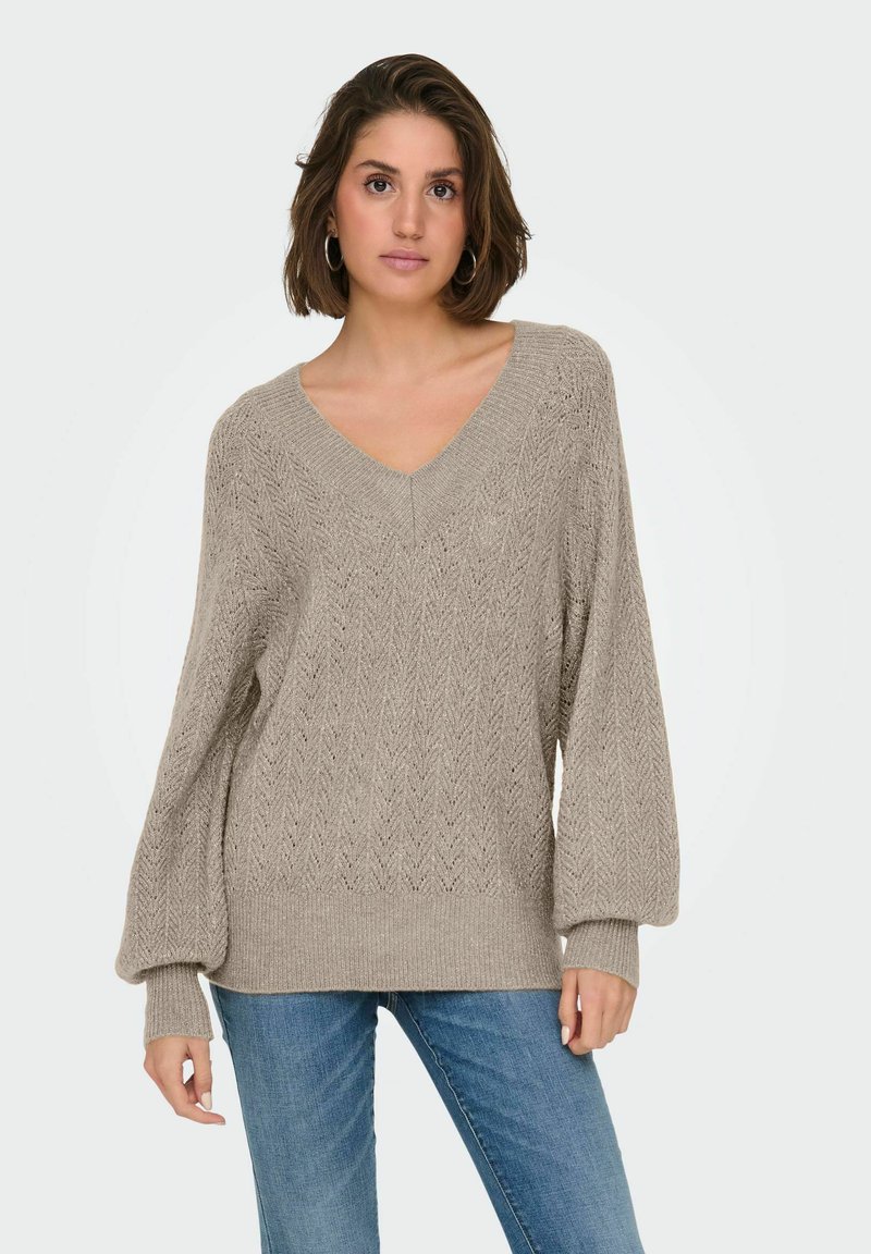 Pull en tricot beige avec un col en V, présentant un motif en chevrons texturé et des manches bouffantes amples. Associé à un jean bleu.