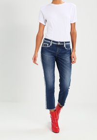 Jeans in denim scuro con bordi sfrangiati, abbinati a una maglietta bianca e stivaletti rossi lucidi alla caviglia. I jeans presentano una vestibilità dritta e tasche laterali.