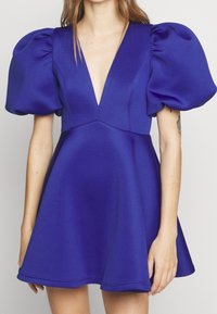 Nainen, joka on pukeutunut lyhyeen, kuninkaallisen siniseen mekkoon, jossa on syvä V-pääntie ja ylisuuria puffihihnoja, seisoo tasaisella taustalla.