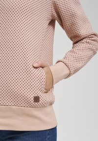 Sudadera de color rosa claro con lunares marrón oscuro, con un bolsillo tipo canguro y puños elásticos. Pequeña etiqueta de marca en el bolsillo.
