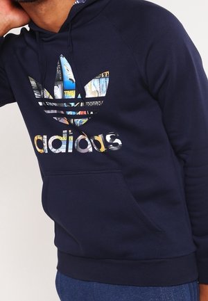 Homme portant un sweat à capuche Adidas bleu marine avec un logo graphique coloré et une poche kangourou à l'avant, main posée sur la nuque.