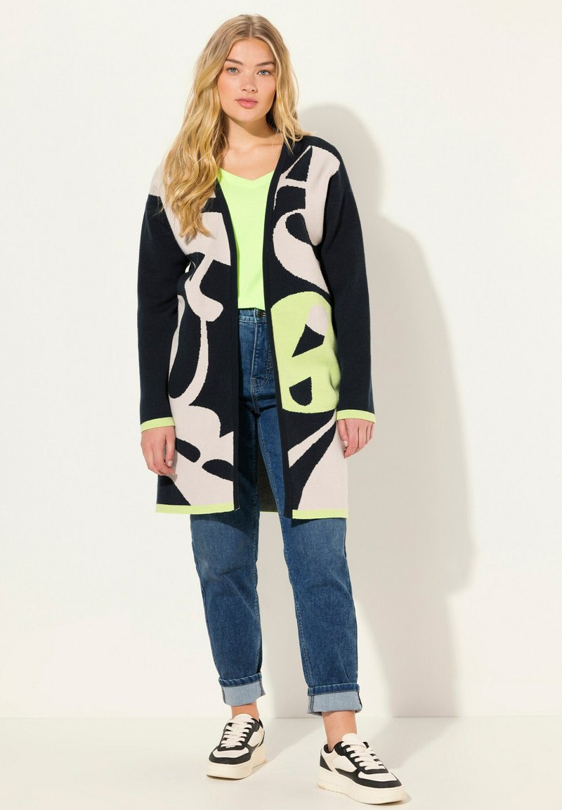 Cardigan lavorato a maglia con motivi astratti in nero, crema e verde, apertura frontale, maniche lunghe, tessuto testurizzato, indossato sopra un top verde neon e jeans.
