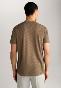Braunes Kurzarm-T-Shirt mit strukturiertem Muster. Rundhalsausschnitt und gerader Saum. Getragen mit hellen Hosen, zeigt die Rückansicht.