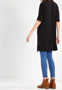 Musta, ylipitkä t-paita, jossa on lyhyet hihat ja sivuhalkiot, päällä siniset tiukat farkut ja ruskeat nilkkurit.