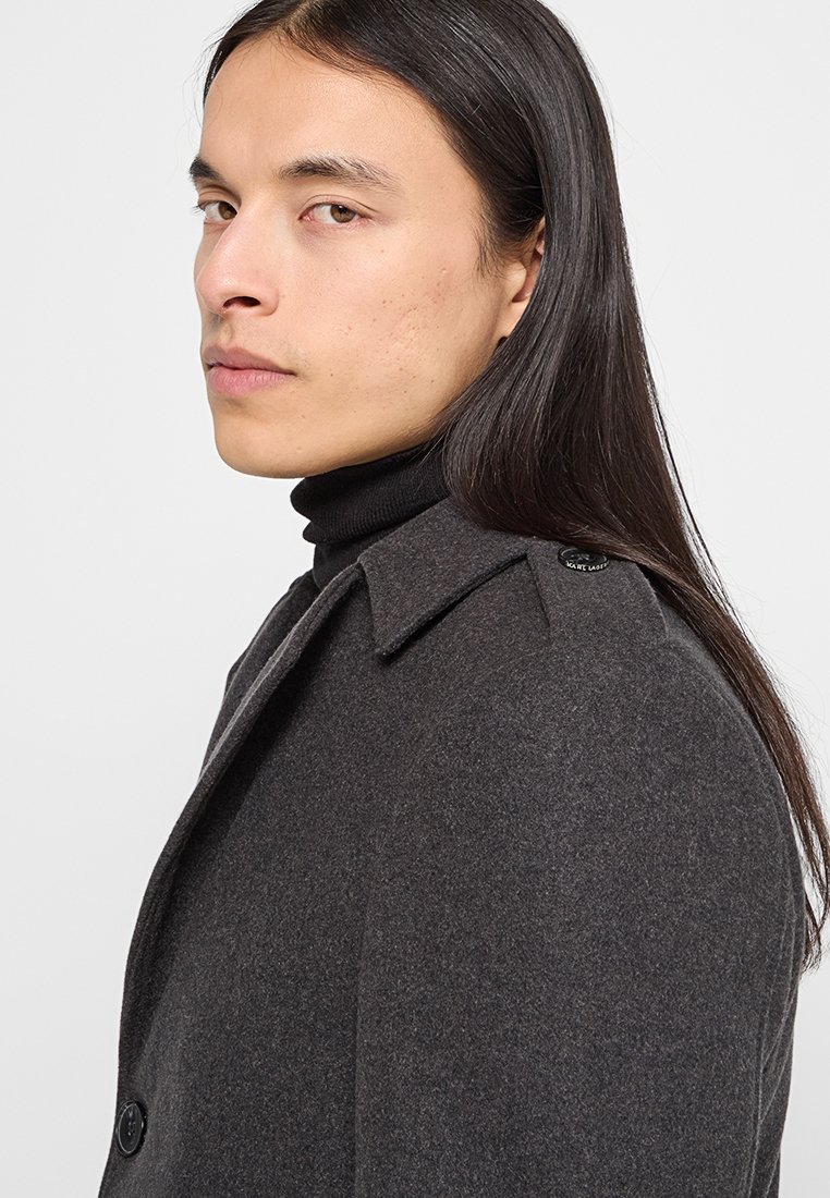 Joven con cabello largo y oscuro que lleva un abrigo gris oscuro y un suéter negro de cuello alto, mirando por encima del hombro frente a un fondo sencillo.