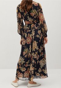 Woman wearing a long-sleeve, ankle-length navy dress with multicolored floral print, tied waist, and white flat shoes, shown from the back.